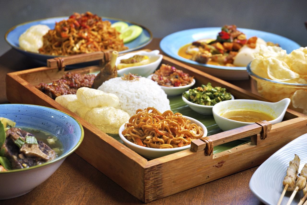 Traditional Indonesian meal with rice, noodles, satay, and various side dishes served on a wooden tray.